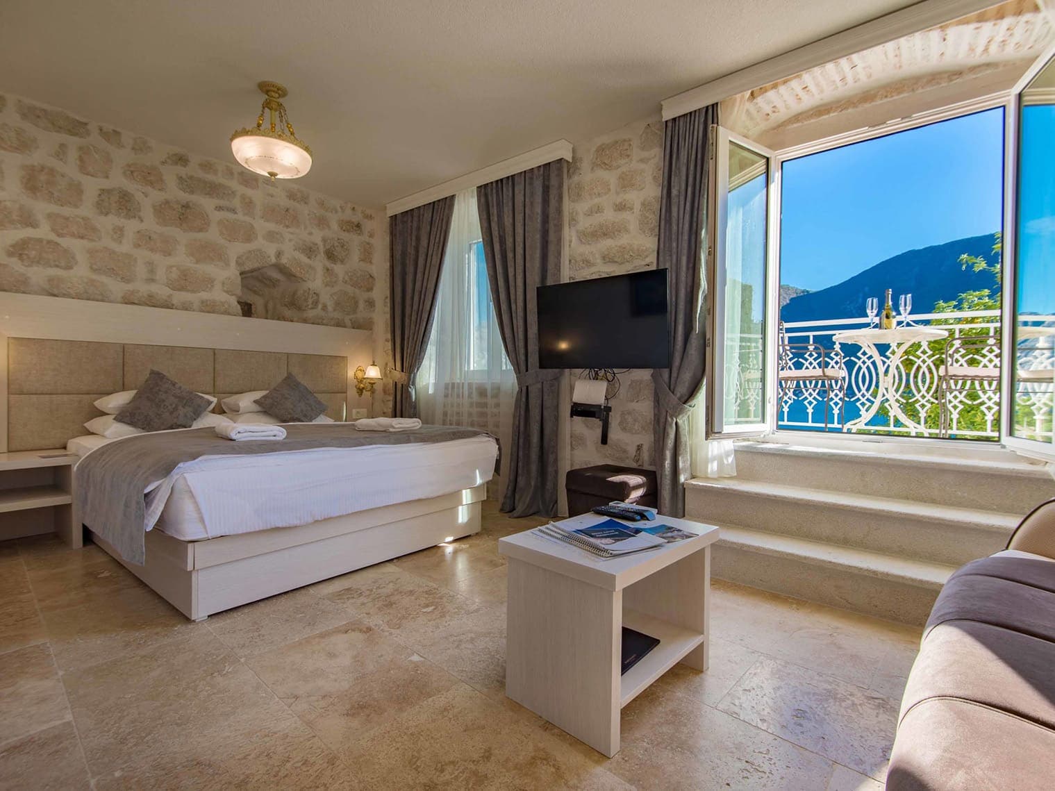 Luxurious hotel room at Hotel Conte featuring a comfortable bed, stone walls, a flat-screen TV, and a balcony with a view of the Bay of Kotor.